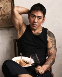Man with tattoos wearing a black tank top holding a wine glass and a plate of food seated indoors.