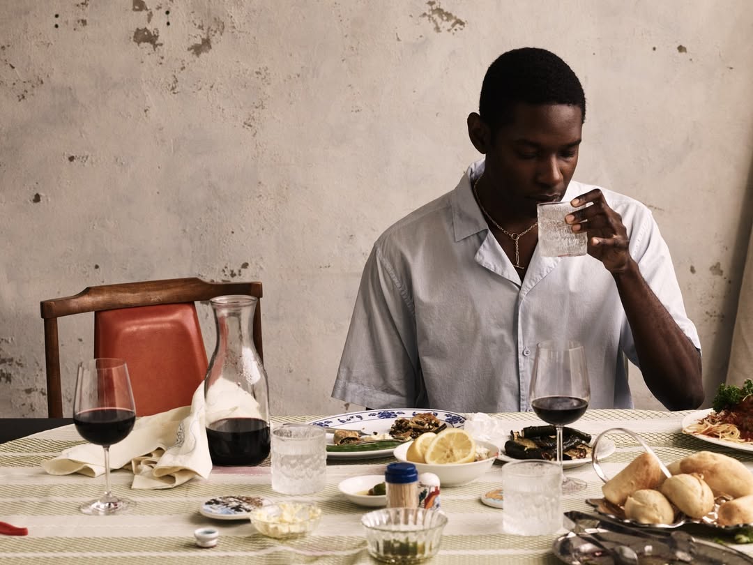 A contemplative dark-skinned man at a table with open light blue shirt, enjoying a meal with wine and water in a cozy, indoor setting.