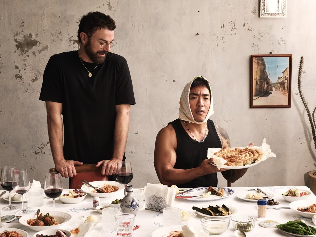 Two men sit at a table laden with food, one in a black t-shirt and glasses stands looking down at the second man who's holding a pie.