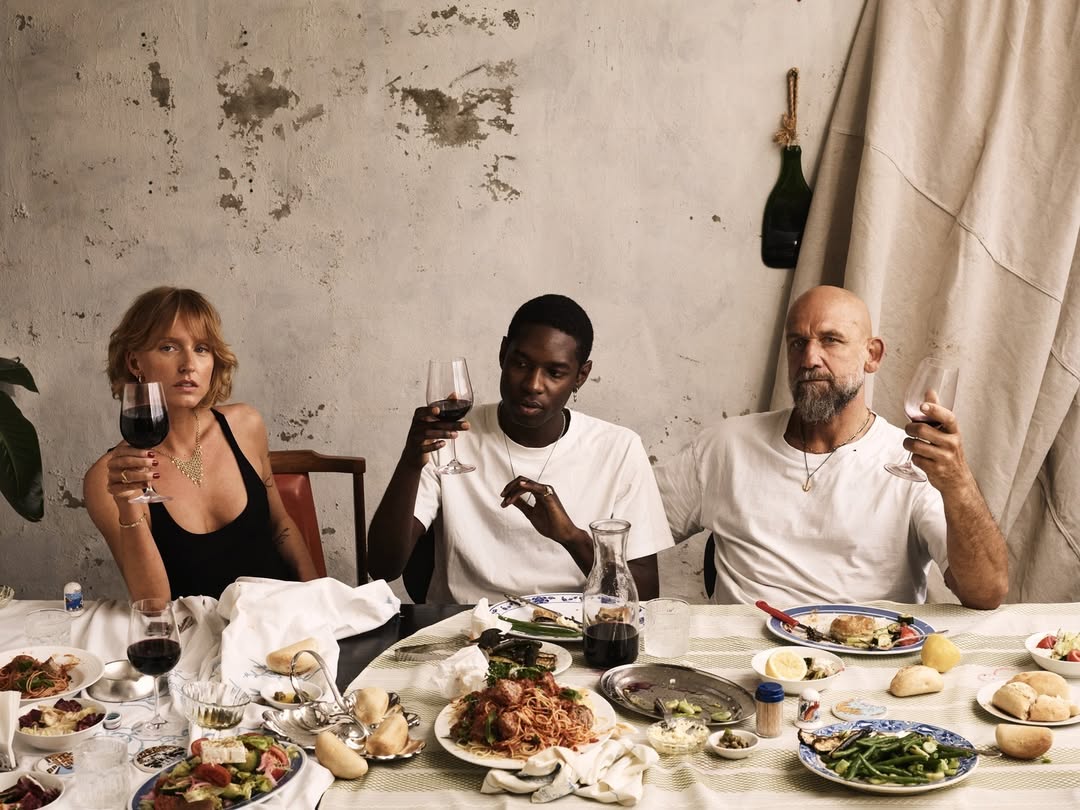 Three people sit at a table covered with food and drinks in an indoor setting with muted colors and natural lighting.