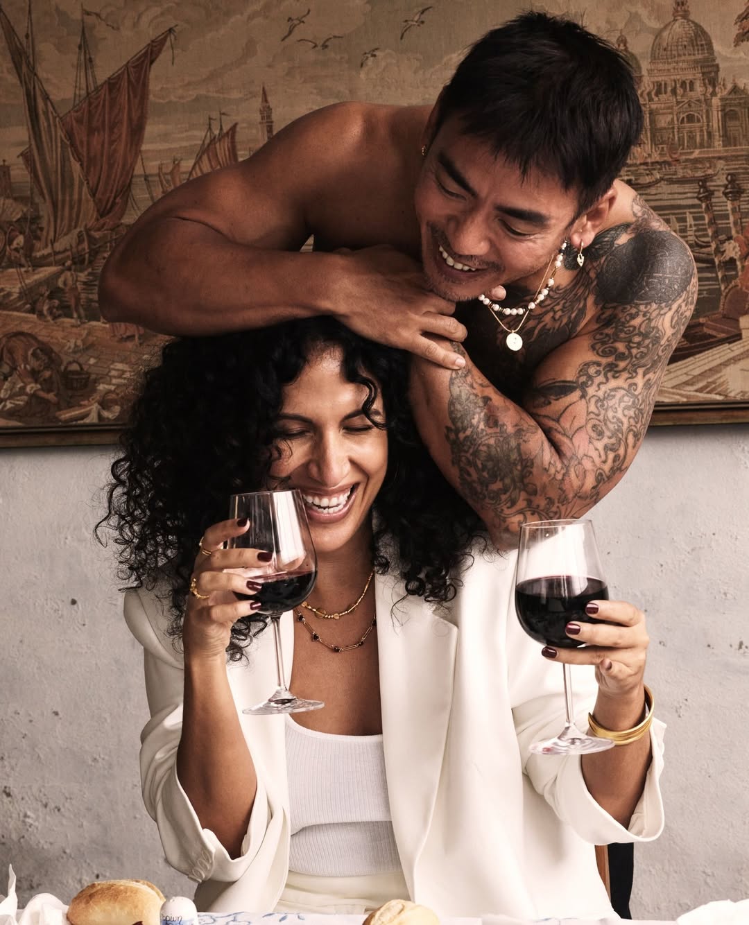 Smiling woman in white blazer holding glass of red wine with muscular man behind in front of painting and table with bread.