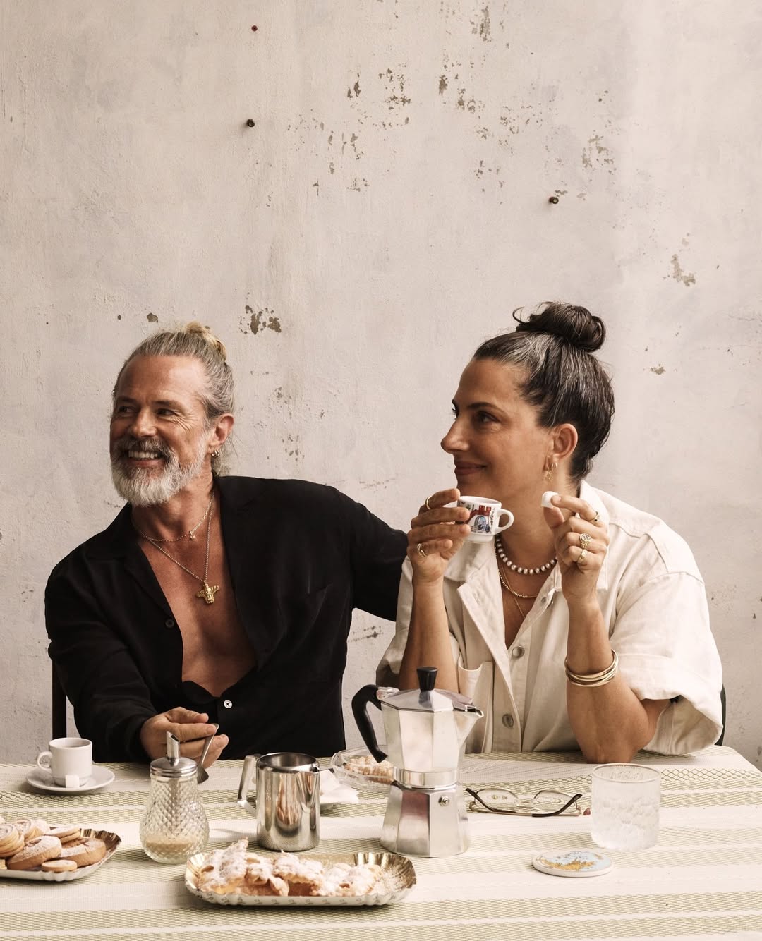 Mature couple sharing coffee and pastry at a table with sunlight, drinks, and tableware in a domestic interior setting.