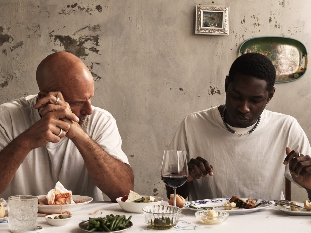 Two men seated at a table with plates of food, in a naturally lit indoor setting with a rustic wall backdrop.
