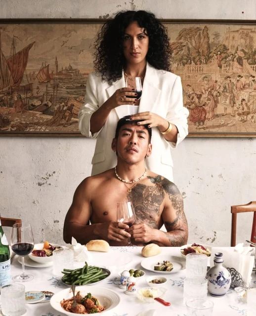 Tattooed man and woman in a white suit with dark curly hair pose indoors in front of a painting over a table of food and drink.