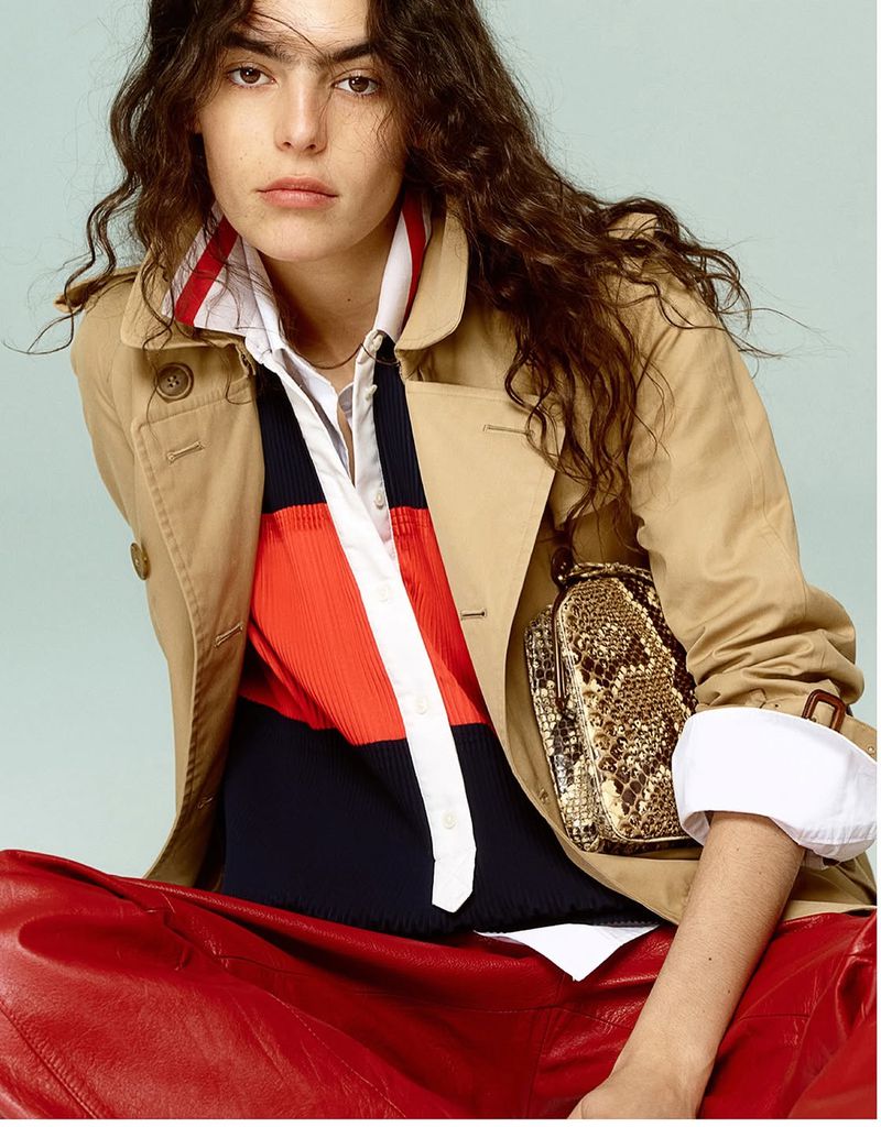 Brunette woman poses in a tan trench coat, red pants, and snakeskin purse against a light blue backdrop for a fashion editorial.