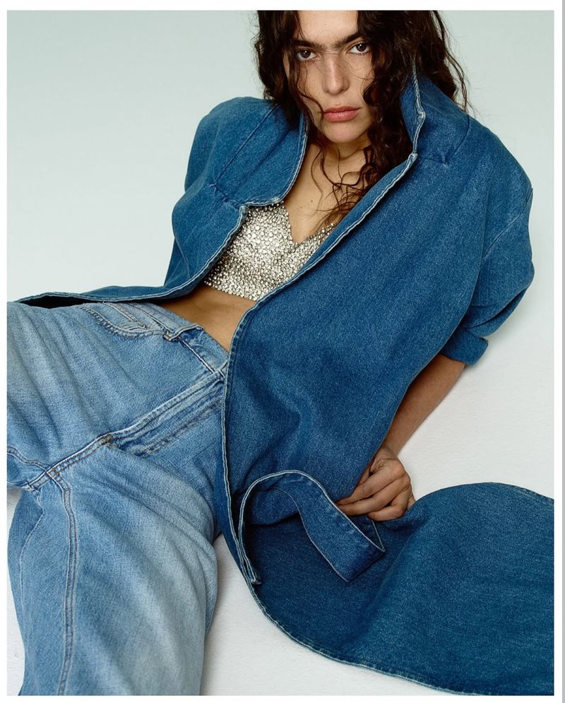 Fashion shot of a woman wearing a denim jacket, jeans, and a sparkly top, posing against a white background in a studio setting.