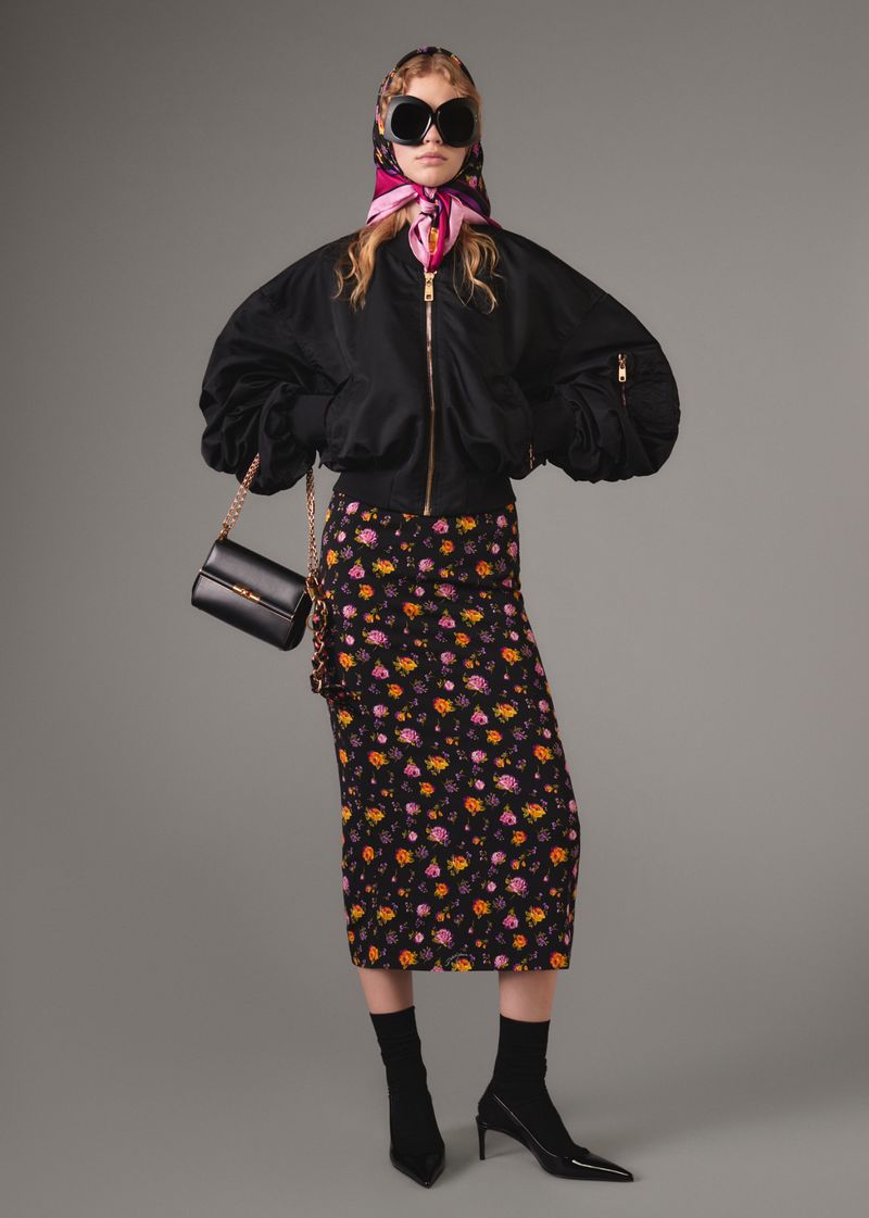 A woman wearing a black bomber jacket, floral skirt, and headscarf poses against a gray background with a black purse.