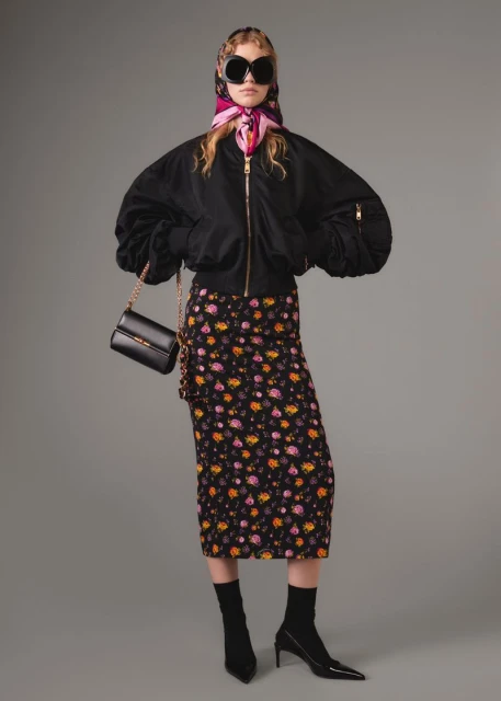 A woman wearing a black bomber jacket, floral skirt, and headscarf poses against a gray background with a black purse.
