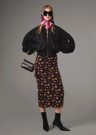 A woman wearing a black bomber jacket, floral skirt, and headscarf poses against a gray background with a black purse.