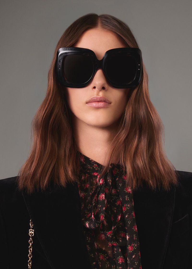A model showcases oversized black sunglasses and a black floral blouse with a black blazer, presenting a high-fashion, modern look.