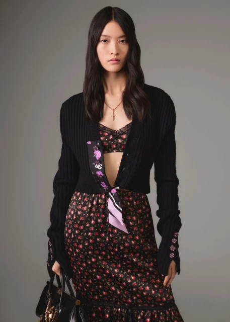 Elegant woman in a black cardigan, floral top and skirt, carrying a black purse, posing in a studio with a neutral background.