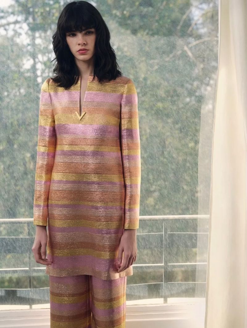 Brunette woman in a striped gold and pink long-sleeve top and pants, standing indoors by a window with a blurred nature background.