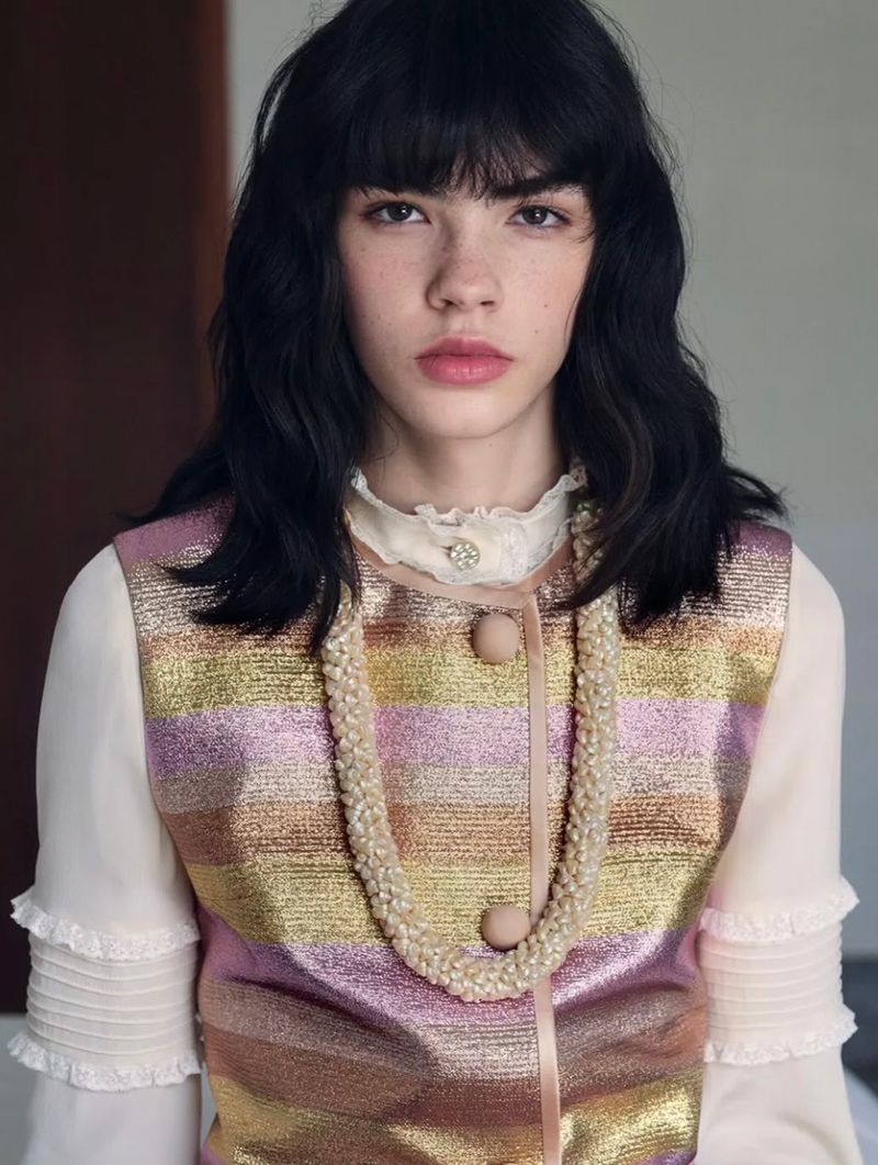 Close-up of a young woman with dark wavy hair wearing a colorful sleeveless top and beaded necklace posing indoors.