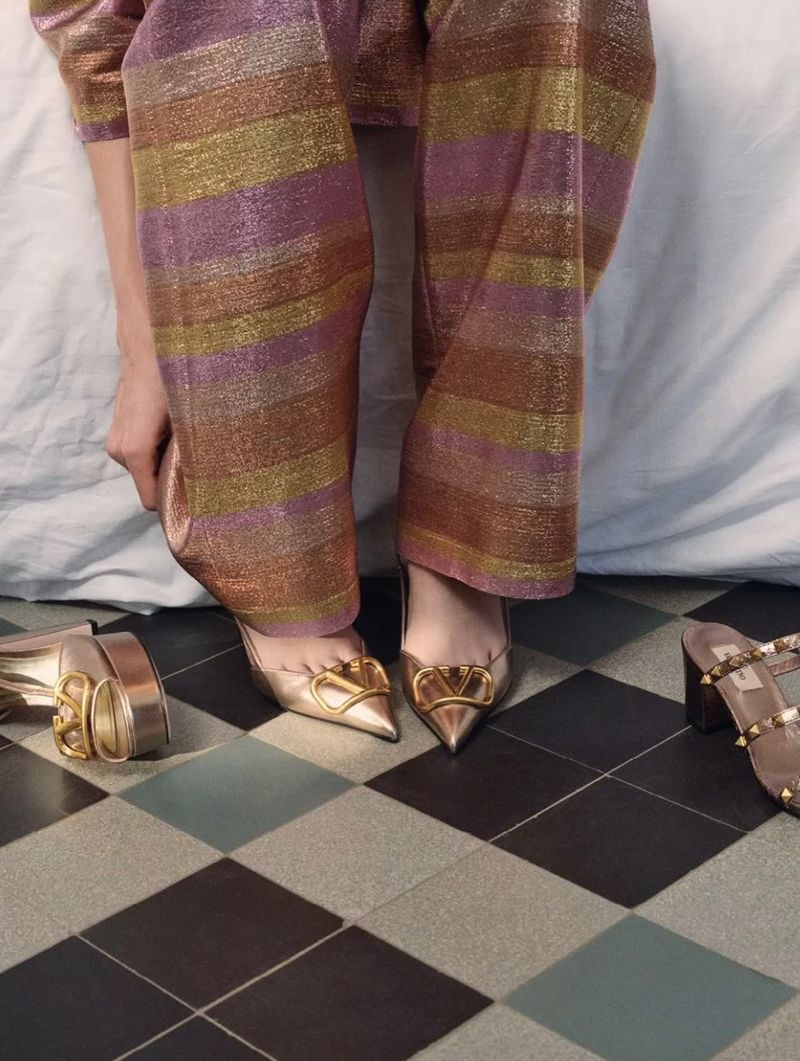 A pair of metallic gold point-toe high heels with hardware details sits on a checkered floor with a woman wearing striped pants.