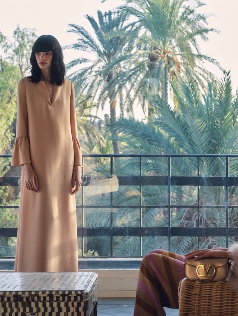 Brunette woman poses in a flowing beige dress on a balcony overlooking palm trees, with a wicker table and purse in the foreground.