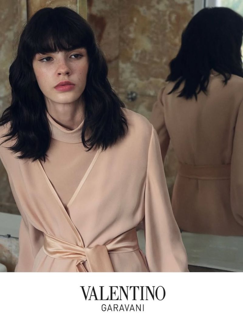 Portrait of a woman with dark hair and bangs wearing a light pink dress with tie detail, reflected in a mirror, fashion ad.