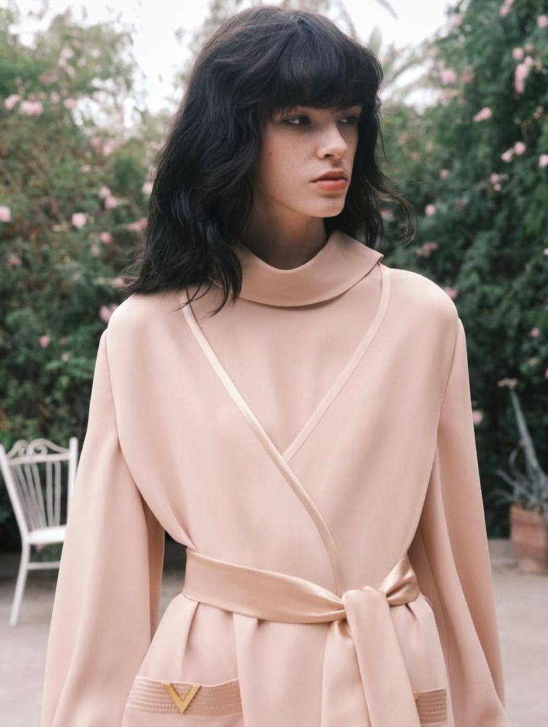 Elegant woman with black hair poses in a peach dress with a belt in an outdoor garden setting with pink flowers and greenery.