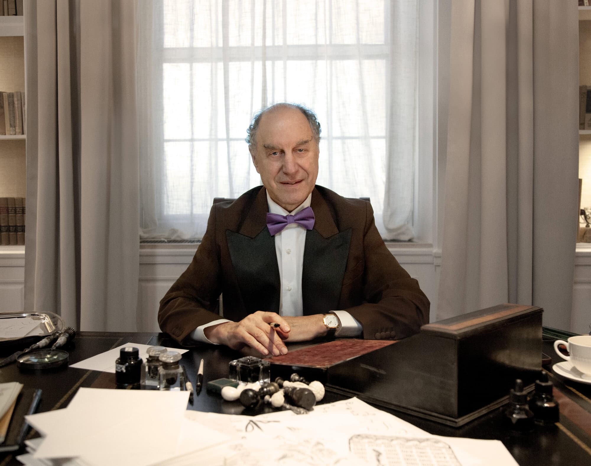 Older man in a brown tuxedo and bow tie sits at a desk with papers, ink, and chess pieces. Light streams through a window behind him.