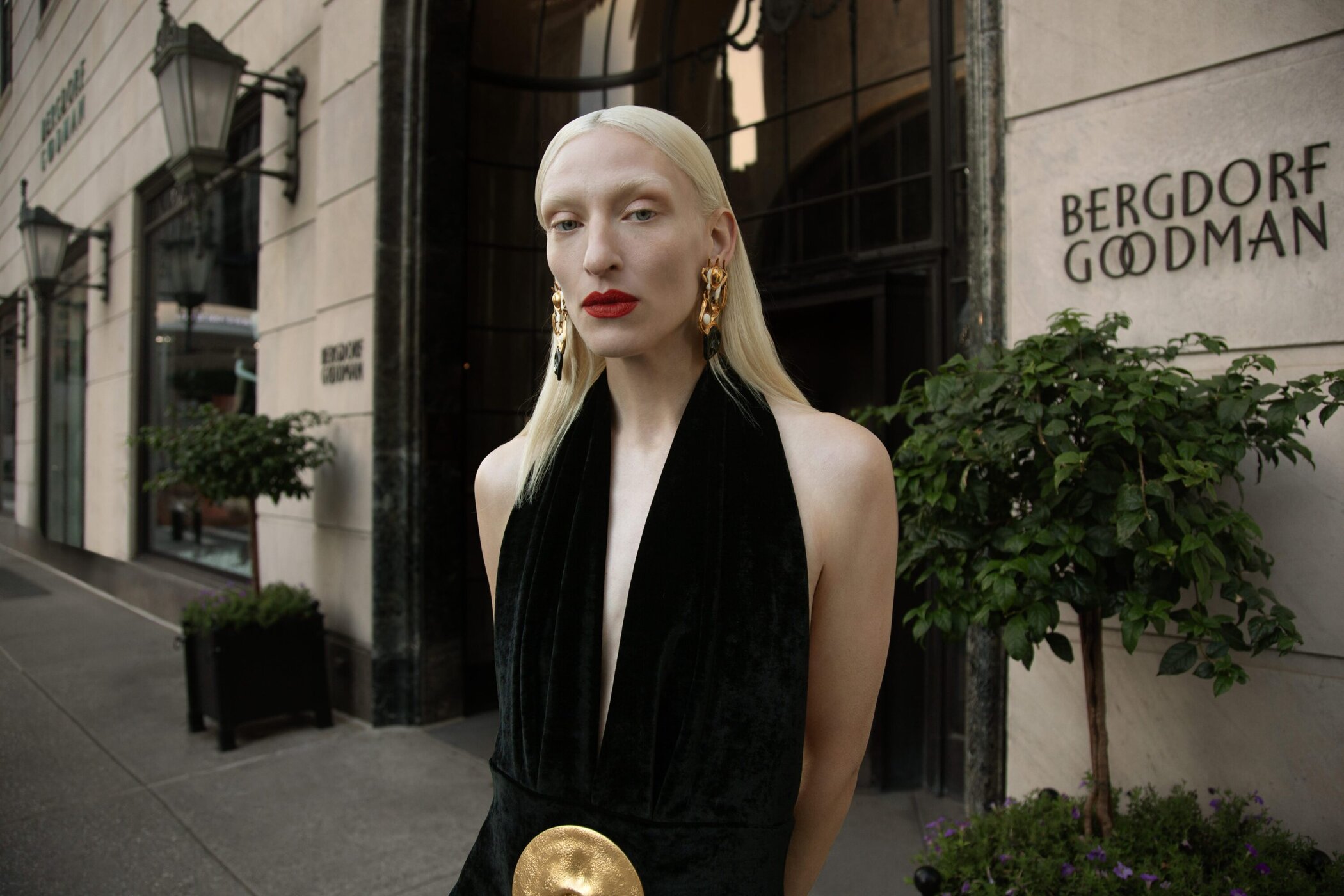 Fashion portrait of a platinum blonde woman wearing a black velvet halter dress with a gold belt posing in front of Bergdorf Goodman.
