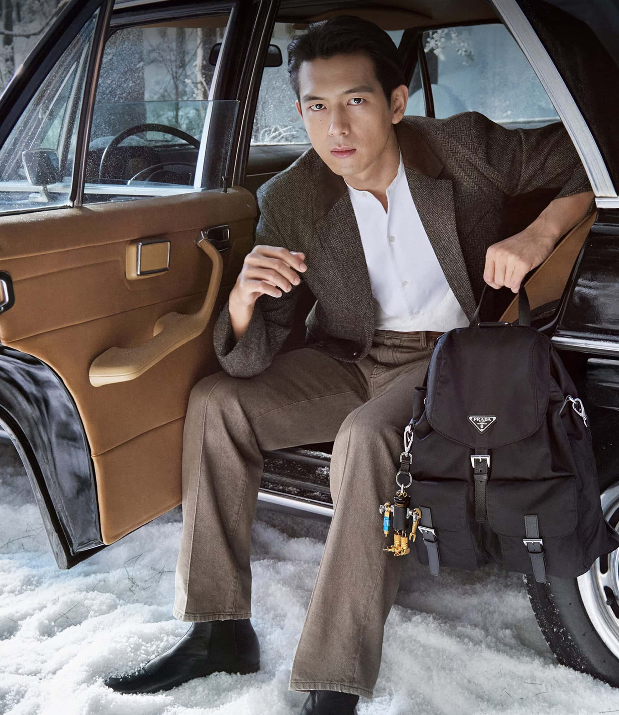 Asian man poses in a tweed suit inside a classic car, holding a backpack and keychain, in an outdoor setting with snow on the ground.