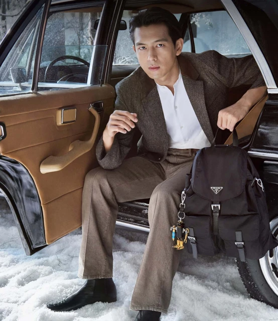 Asian man poses in a tweed suit inside a classic car, holding a backpack and keychain, in an outdoor setting with snow on the ground.