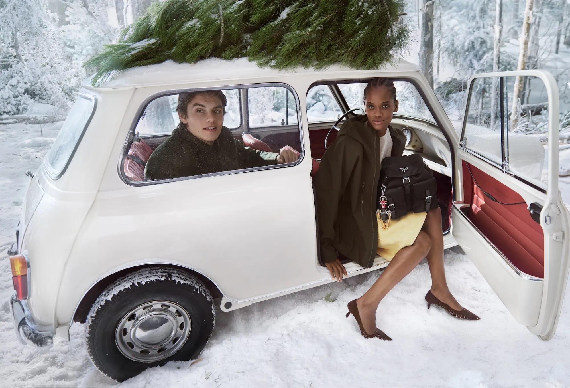 Two people with a white vintage car in the snow, adorned with a Christmas tree on top, one woman getting out of the car with Prada bag.