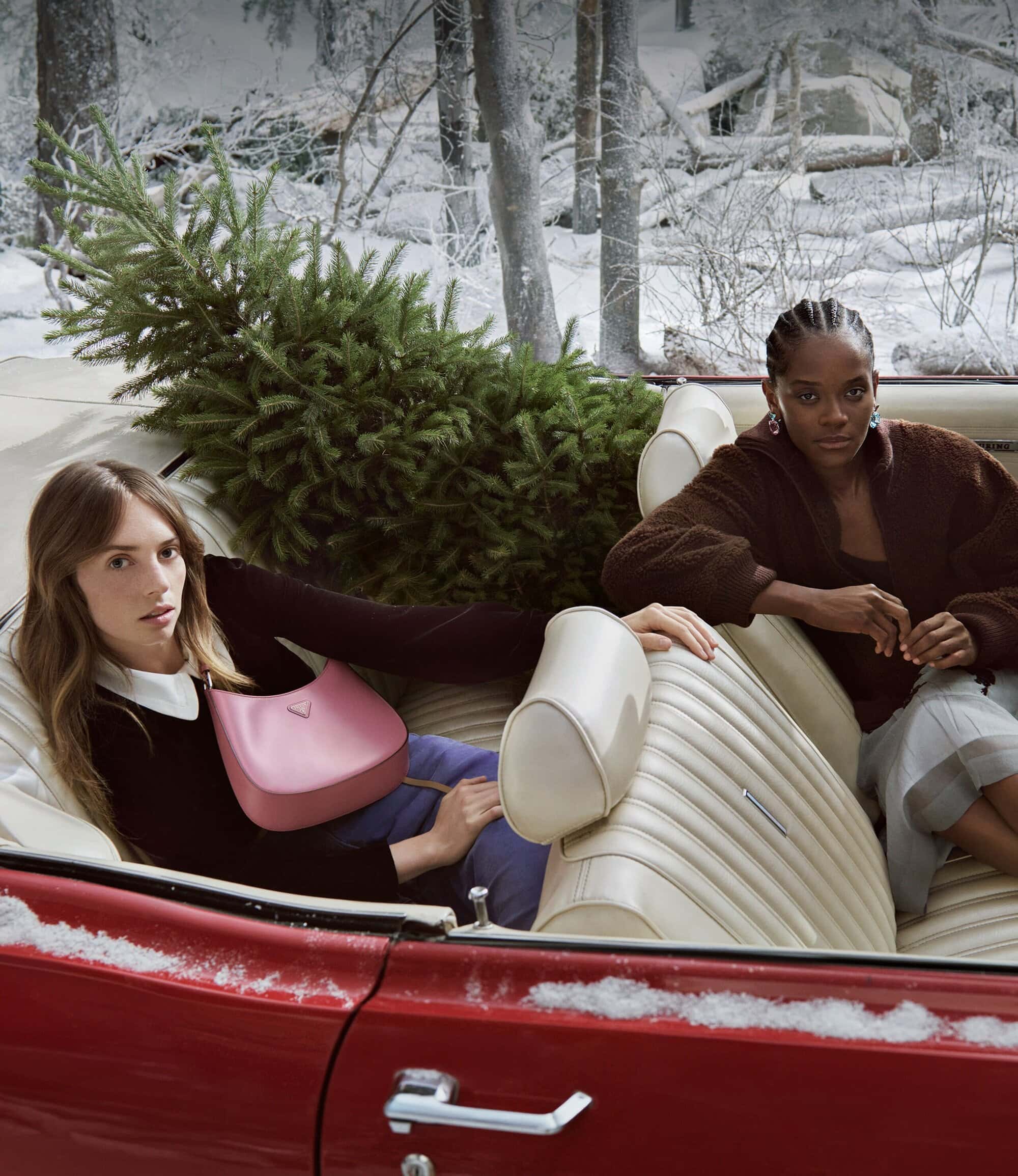 Two women pose in a red convertible with a Christmas tree against a wintry forest backdrop, featuring a pink handbag.
