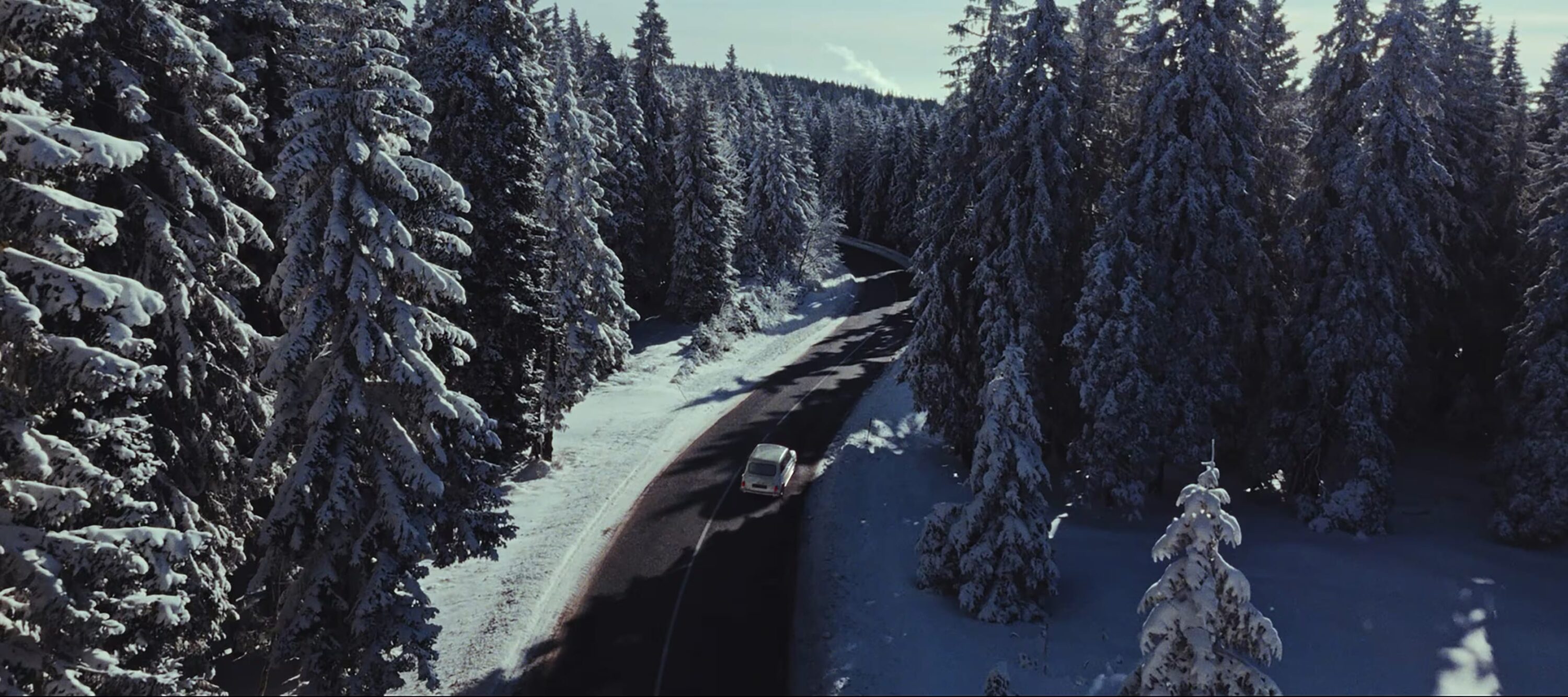 Aerial view of a white car driving on a paved road through a dense forest covered in snow on a sunny winter day.