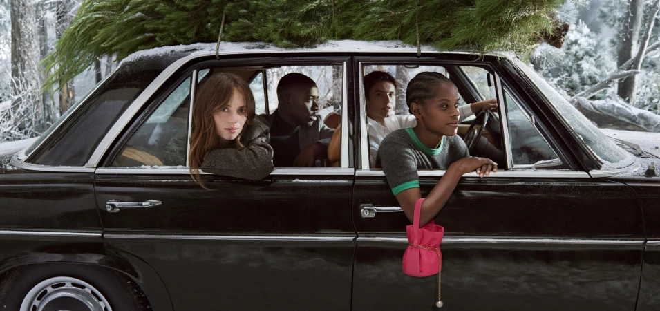 Group of four people inside a vintage car in a wintery forest with a Christmas tree tied to the roof, showcasing a pink purse.