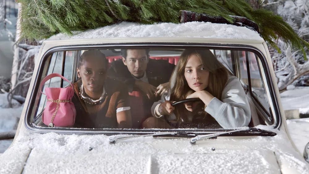 Three people inside a snow-covered car with a tree on top. The car sits in a snowy environment with bare trees and light-colored buildings.