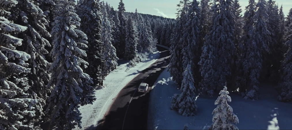 Aerial view of a car driving along a winding asphalt road through a dense, snow-covered pine forest.