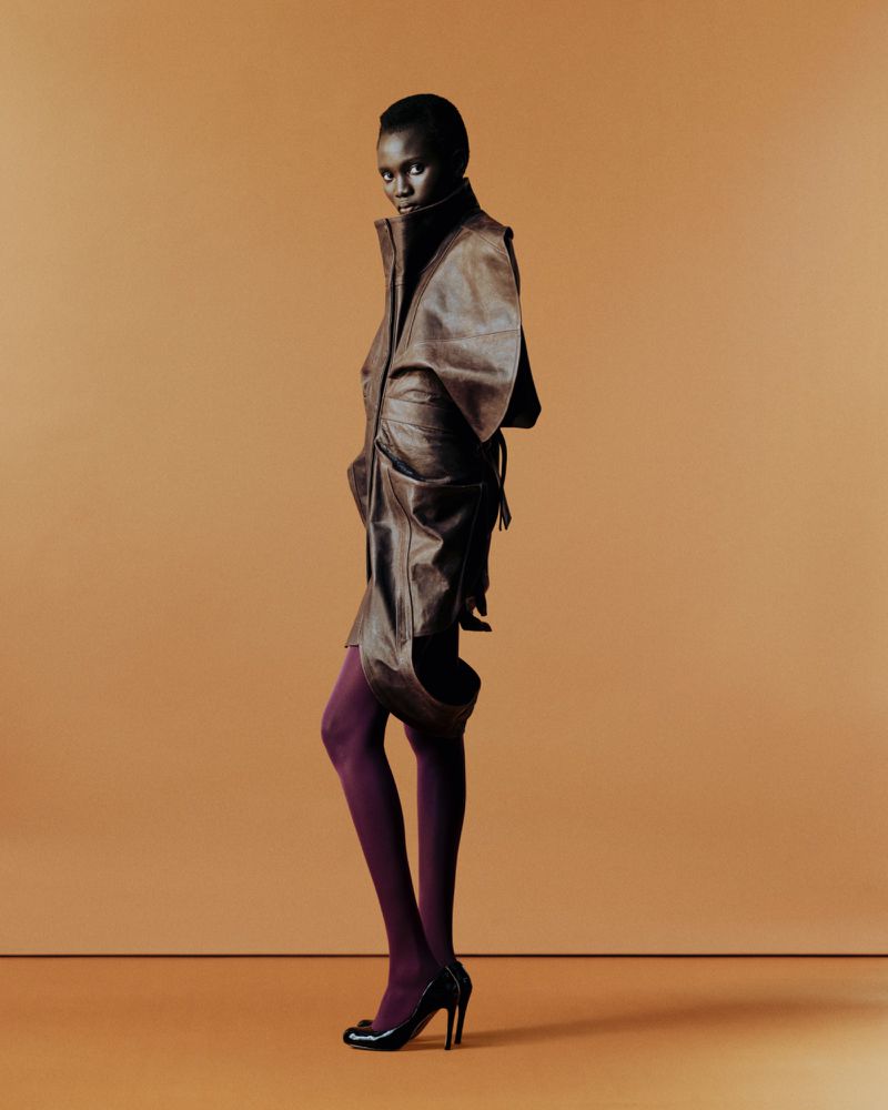 Full-length shot of an African woman in a brown leather outfit and purple tights, posing against a tan studio backdrop in high heels.