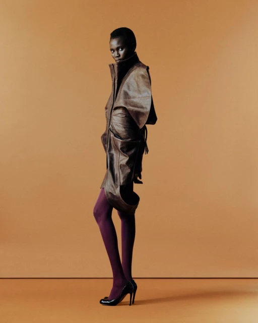 Full-length shot of an African woman in a brown leather outfit and purple tights, posing against a tan studio backdrop in high heels.