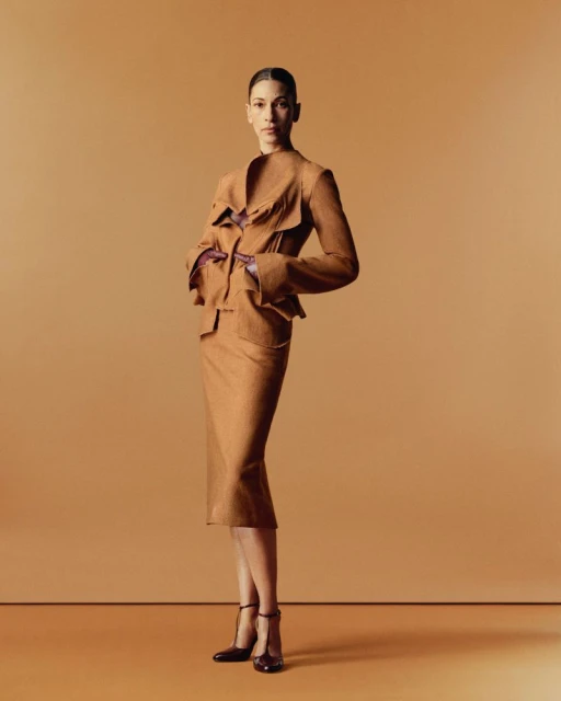 A woman in a tan blazer and pencil skirt poses in a studio with brown monochrome background and black heels, emphasizing a fashion editorial.