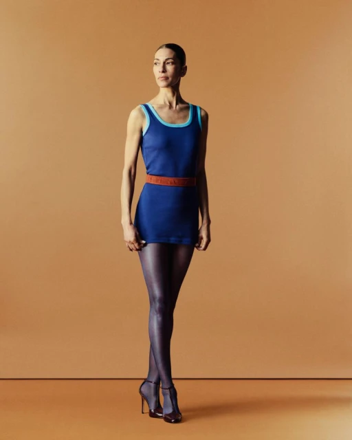 Full-length portrait of a woman with fair skin in a blue dress with black tights and high heels posing in front of a tan backdrop.