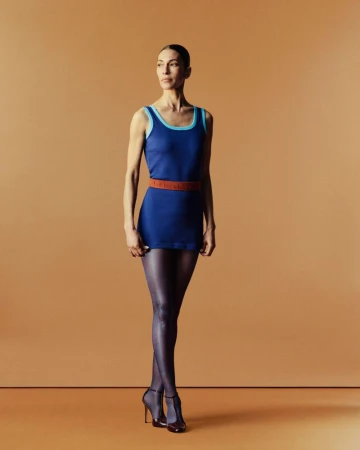 Full-length portrait of a woman with fair skin in a blue dress with black tights and high heels posing in front of a tan backdrop.