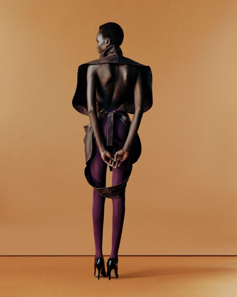 Fashion portrait from the back of a Black female adult wearing a sculptural garment, purple tights, and high-heeled shoes on an orange backdrop.