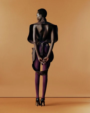 Fashion portrait from the back of a Black female adult wearing a sculptural garment, purple tights, and high-heeled shoes on an orange backdrop.