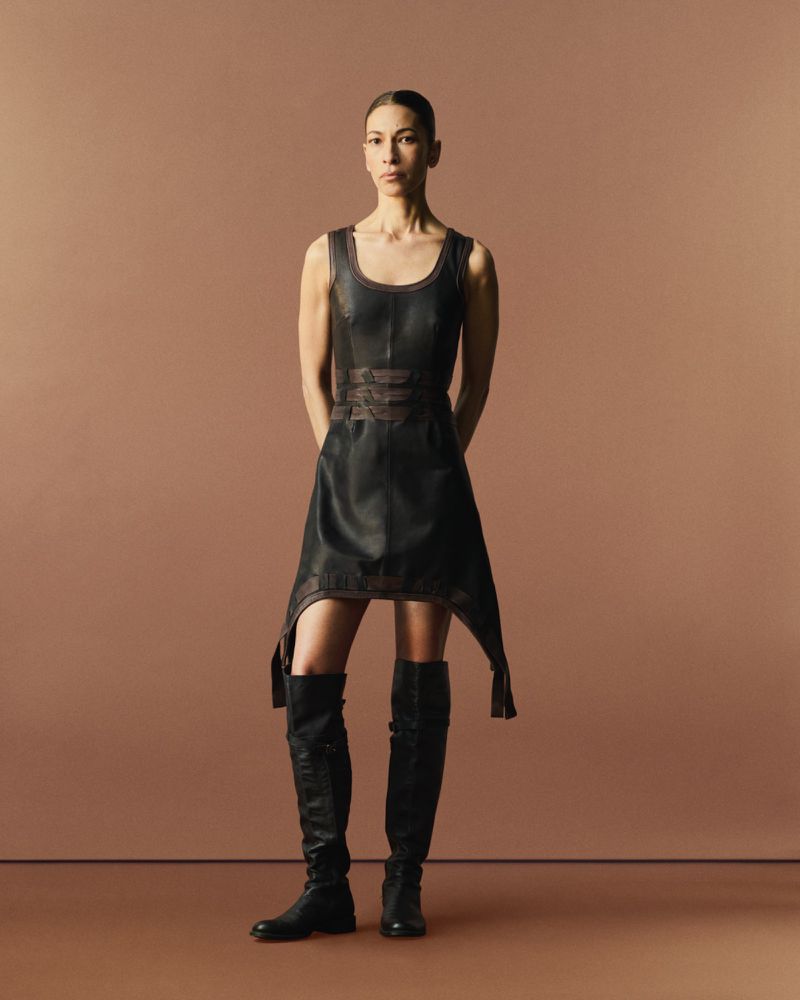 A woman in a black dress and boots stands in a studio. The black dress has an asymmetrical cut and the boots are black.