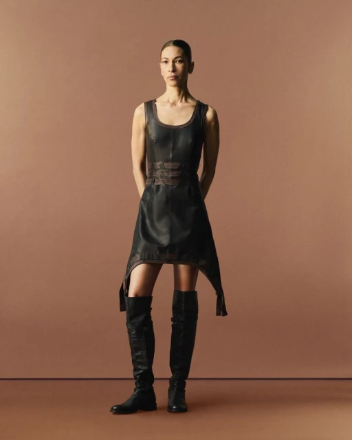 A woman in a black dress and boots stands in a studio. The black dress has an asymmetrical cut and the boots are black.