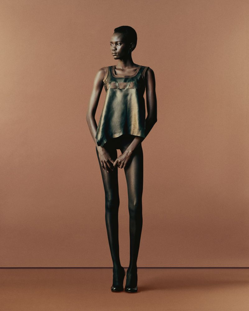 Full-length studio shot of a slender Black woman in a shiny tank top, dark tights, and high heels against a warm brown background.