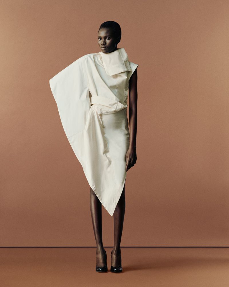 A single Black female fashion model in a draped white dress poses against a monochromatic tan background with dark hair and red heels.