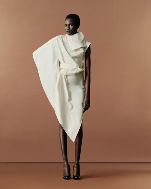 A single Black female fashion model in a draped white dress poses against a monochromatic tan background with dark hair and red heels.