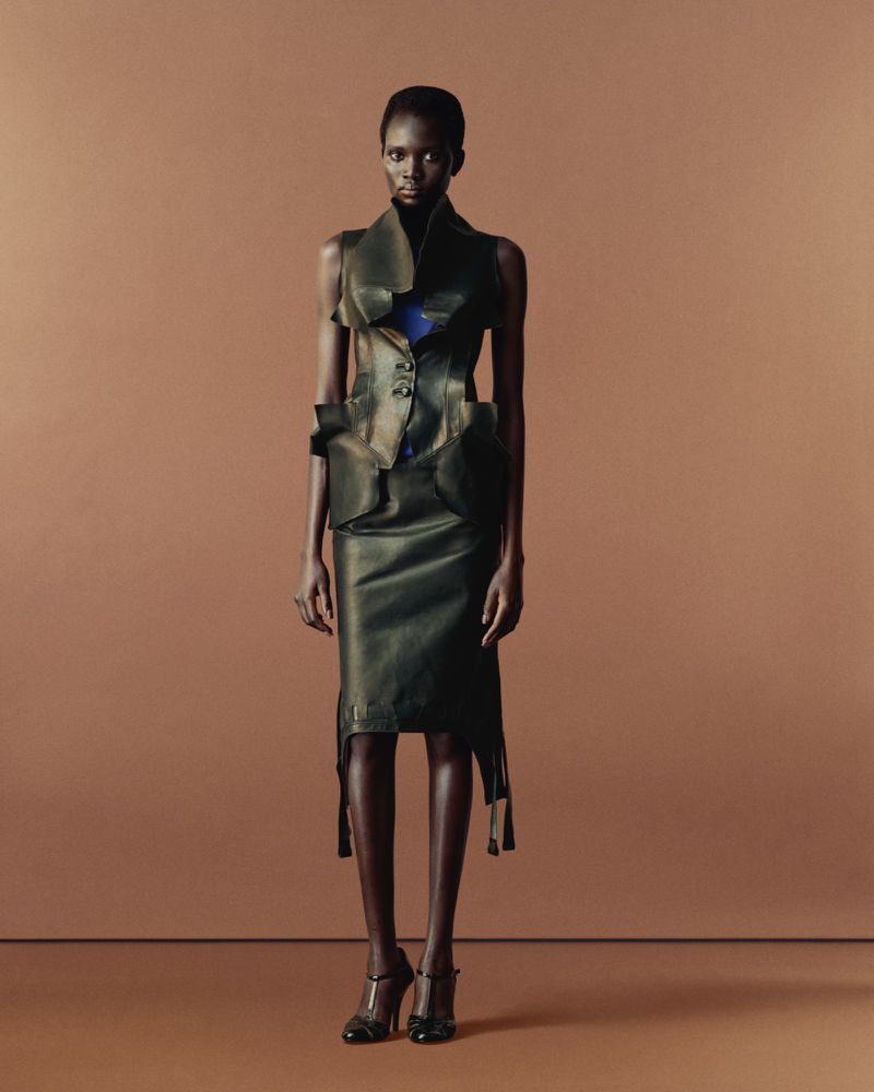 A slender African woman in an avant-garde black leather outfit with a sleeveless vest and skirt stands on a tan background, fashion forward.