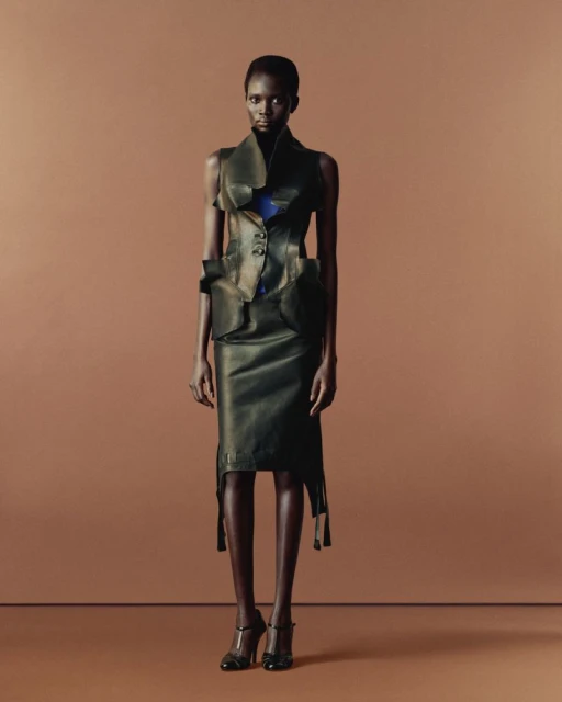A slender African woman in an avant-garde black leather outfit with a sleeveless vest and skirt stands on a tan background, fashion forward.