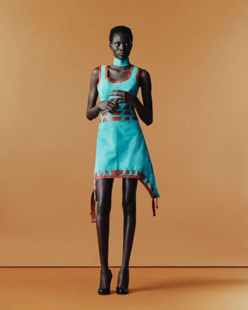 A striking fashion portrait of an adult female model with dark skin in a sleeveless teal dress and black high heels against tan backdrop.
