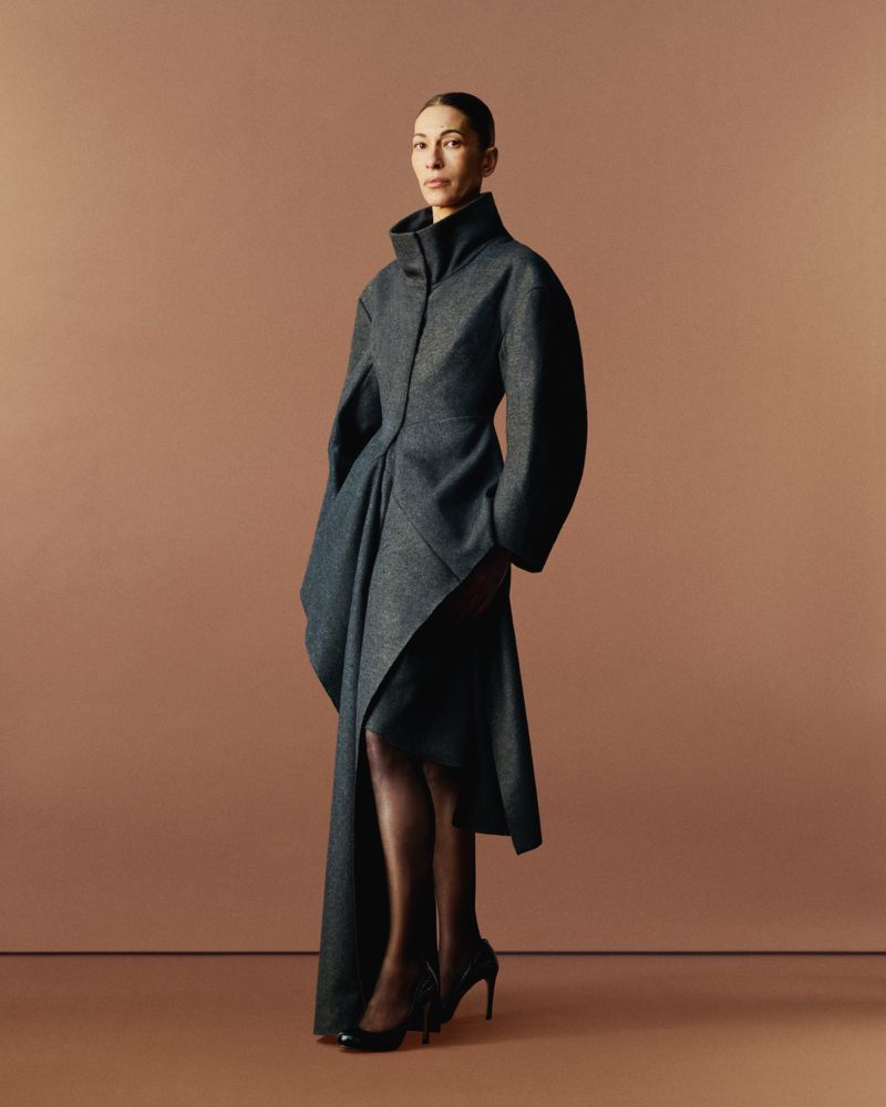 Elegant adult female poses in studio wearing a full-length grey wool coat with black heels and dark stockings against a light brown backdrop.