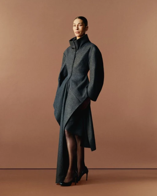 Elegant adult female poses in studio wearing a full-length grey wool coat with black heels and dark stockings against a light brown backdrop.