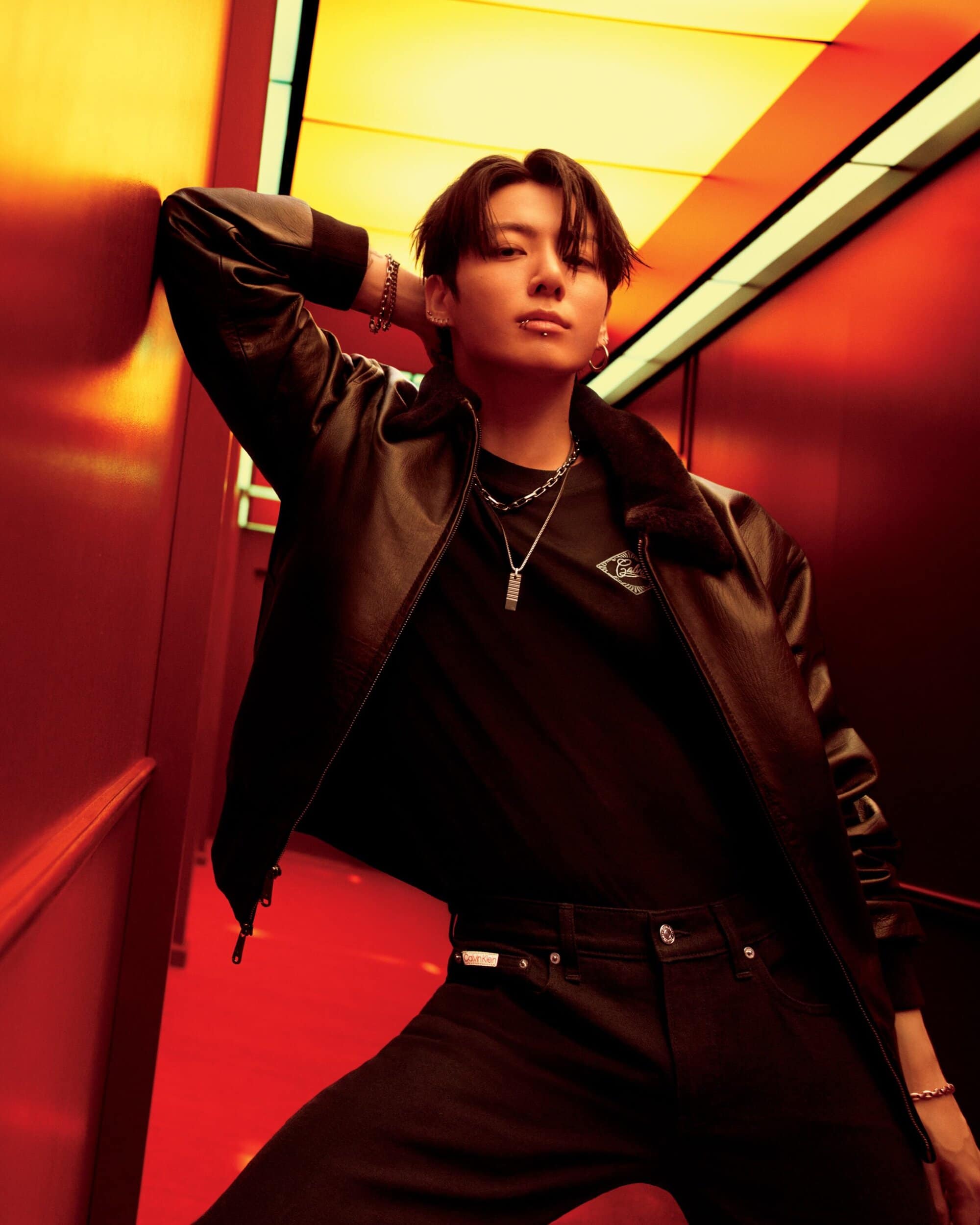 Jungkook from BTS poses in a black leather jacket and black jeans, set against a vibrant red and yellow tunnel corridor.