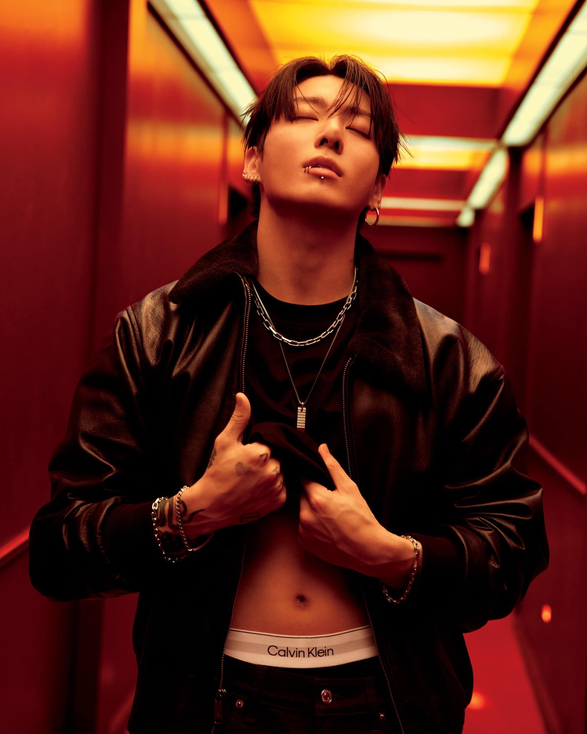 Adult man with lip piercing poses in a black leather jacket and jeans, lifting his black t-shirt to reveal underwear in a studio.